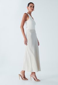 Hame maxi berbahan beige yang mengalir dengan tekstur halus, dipadukan dengan atasan tank top putih yang berdempul serta sandal hak tinggi nude.