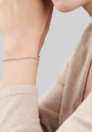 Zarter silberner Kettenarmband an einem Handgelenk, getragen von einer Person in einem beigen Langarmshirt, vor einem schlichten hellgrauen Hintergrund.