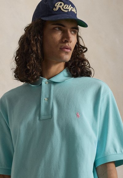 Junger Mann mit langen lockigen Haaren, der eine blaue Ralph-Baseballkappe und ein hellblaues Polohemd mit einem pink bestickten Logo trägt.