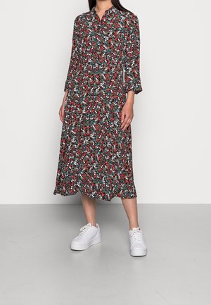 Frau trägt ein midiklanges Blumenkleid mit roten, weißen und grünen Mustern, kombiniert mit weißen Plateausneakern, und steht vor einem schlichten Hintergrund.