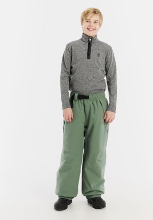 Grijze lange mouwen top met een zwarte rits en logo, gecombineerd met losse groene broek met een riem en zijzakken. Zwarte schoenen maken de look compleet.