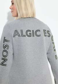 Sudadera gris de manga larga, con texto oscuro en negrita en la parte posterior. El tejido parece suave y de un ajuste relajado. Sin patrones adicionales.