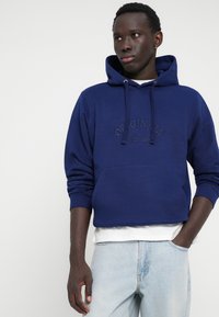 Sweat-shirt à capuche en coton bleu marine avec poche avant, logo "ORIGINALS" en relief, poignets côtelés et coupe décontractée. Porté sur une chemise blanche.