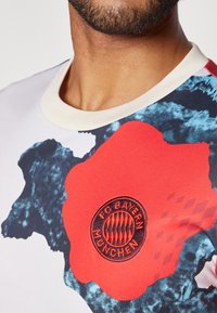 Lyhythihainen t-paita valkoisena ja sinisenä, jossa on suuri punainen graafinen painatus ja mustalla brodeeratussa FC Bayern München -logossa rintamalla.