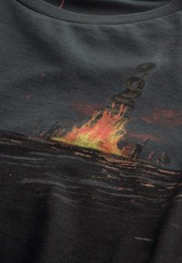 T-shirt nera in cotone con una grafica che rappresenta un incendio su una piattaforma petrolifera, con fiamme arancioni e gialle su uno sfondo scuro.