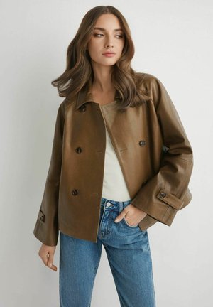 Kobieta o falistych brązowych włosach, ubrana w brązową, dwurzędową skórzaną kurtkę, biały top i niebieskie dżinsy, stojąca na tle jednolitego jasnego tła.