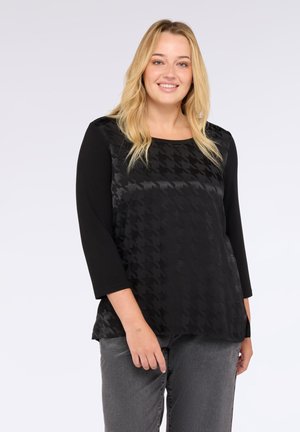 Femeie zâmbitoare cu păr blond poartă un top negru cu model, cu mâneci până la trei sferturi, și blugi gri închis, pe un fond simplu.