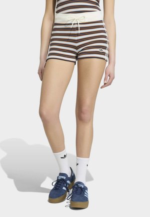 Žena u prugastim pletenim kratkim hlačama s vezicama, bijelim Adidas čarapama i plavim Adidas tenisicama na jednobojnoj svijetloj pozadini.