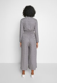 Mono de tela estampada, con mangas largas y cintura fruncida, y pantalones de pierna ancha. Los colores incluyen azul marino y crema.