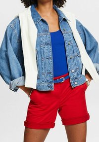Personne portant un débardeur bleu, une veste en jean aux manches retroussées, un short rouge avec revers, une ceinture bleue et un pull blanc drapé sur les épaules.
