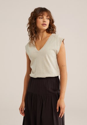 Frau mit welligem Haar, die ein hellgraues ärmelloses V-Ausschnitt-Top und einen hoch taillierten schwarzen Rock vor neutralem Hintergrund trägt.