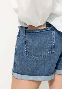 Shorts en denim avec un délavage bleu, ourlet retroussé, poche arrière et coutures visibles. Associés à une chemise légère et fluide.