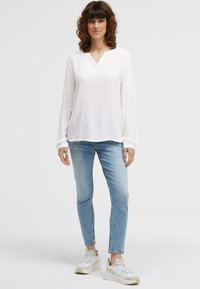 Frau mit lockigem braunem Haar, die eine weiße Bluse mit langen Ärmeln, hellblaue Jeans und weiße Sneakers trägt, steht vor einem schlichten Hintergrund.