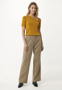 Femme portant un haut à manches courtes moutarde, un pantalon large à carreaux marron taille haute et des bottines noires se tenant contre un fond blanc.