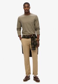 Beżowa koszula z długim rękawem, khaki spodnie, koszula w kratę zawiązana w talii, brązowe skórzane buty z sznurowadłami i ozdobnym przeszyciem, zegarek na nadgarstku.