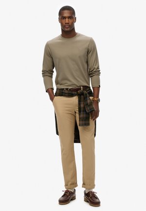 Beige longslavenshirt, kaki broek, geruite shirt om de taille geknoopt, bruine leren schoenen met veters en decoratieve stiksels, horloge om de pols.