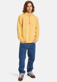 Sudadera amarilla con un logo, bolsillo frontal y cordón. Combinada con jeans azules holgados y zapatillas marrones.