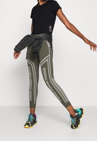 Musta lyhythihainen paita, joka on yhdistetty oliivinvihreisiin legginsseihin, joissa on harmaita yksityiskohtia ja verkkoaineksia, ja asusteena mustat lenkkarit keltaisilla pohjilla.