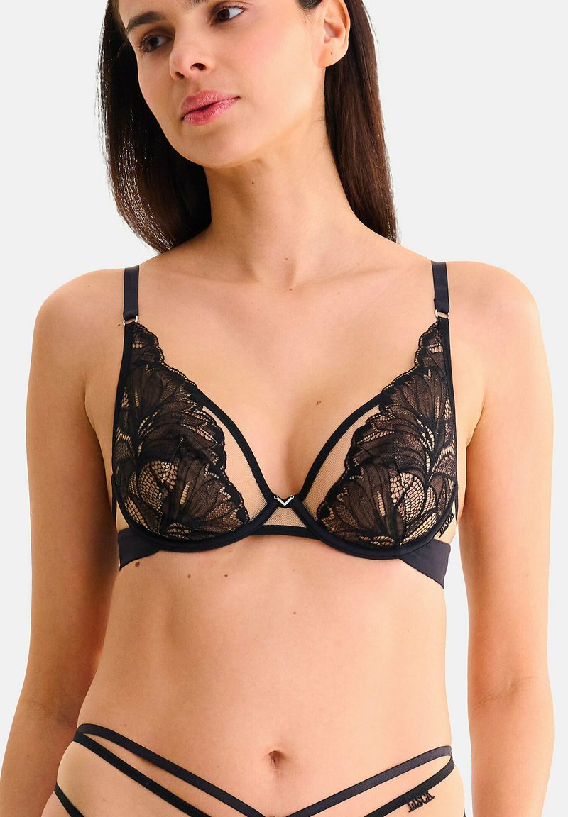 Femme portant un soutien-gorge en dentelle noire avec armatures et un bas assorti à lanières, faisant face légèrement à gauche avec une expression neutre, sur fond uni.