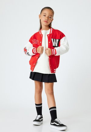 Giacca varsity rossa e bianca con una lettera ricamata, indossata sopra un maglione bianco e una gonna nera, abbinata a calze al ginocchio e sneakers nere.