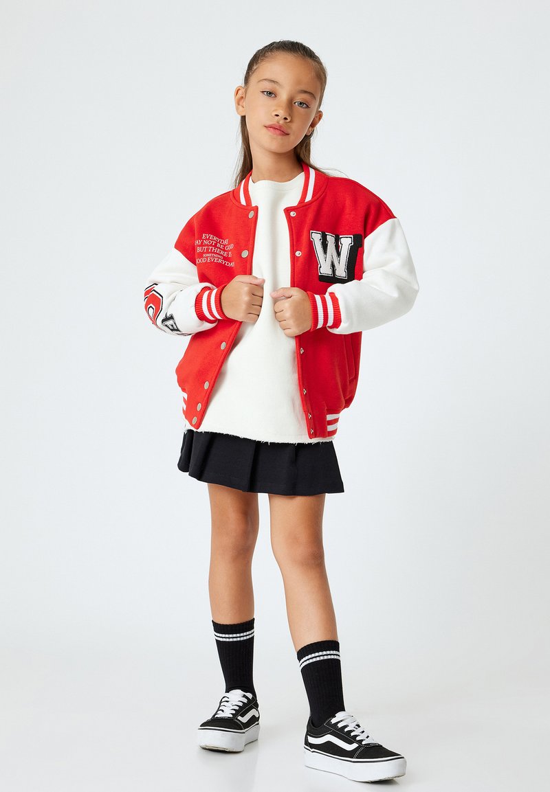 Rot-weiße Collegejacke mit gesticktem Buchstaben, getragen über einem weißen Pullover und einem schwarzen Rock, kombiniert mit kniehohen Socken und schwarzen Turnschuhen.