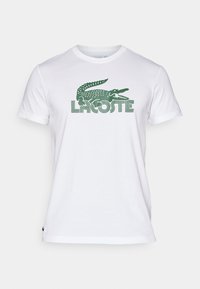 Camiseta de algodón blanca con un logo de cocodrilo verde y el texto "LACOSTE". Ajuste clásico, cuello redondo y mangas cortas. Textura suave.