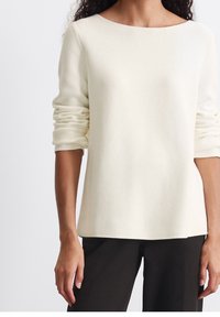 Femme portant un pull crème à manches longues froncées et un pantalon noir, debout devant un fond blanc uni.