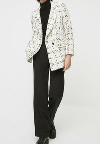 Blazer strutturato bianco con motivo a quadri neri, chiusura con un bottone e due tasche anteriori, abbinato a pantaloni larghi neri.