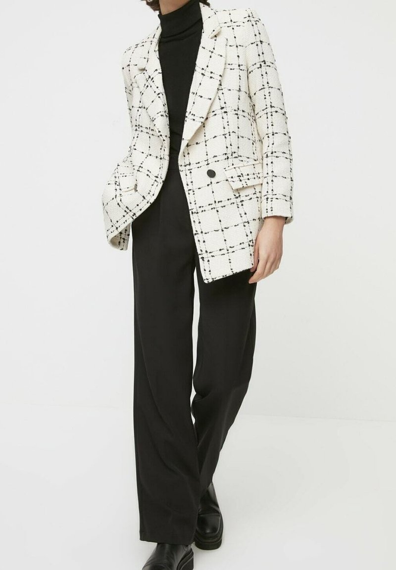 Blazer strutturato bianco con motivo a quadri neri, chiusura con un bottone e due tasche anteriori, abbinato a pantaloni larghi neri.
