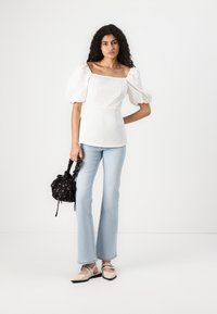 Bluză albă cu mâneci bufante și decolteu pătrat, asortată cu blugi evazați de culoare albastru deschis. Accesoriile includ o geantă neagră cu un model și pantofi crem.
