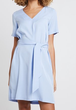 Vrouw draagt een lichtblauwe jurk met korte mouwen, een V-hals en een brede stoffen riem die in de taille is gestrikt.