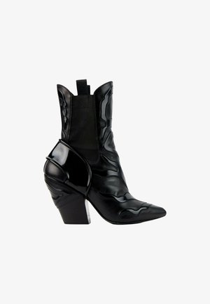Bottine en cuir noir à bout pointu, talon épais, panneaux latéraux élastiques et motifs décoratifs en relief sur fond blanc.
