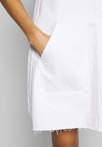 Bílé džínové šaty se dvěma velkými předními kapsami, třepeným lemem a jednoduchým designem. Materiál se zdá být pevný a mírně texturovaný.