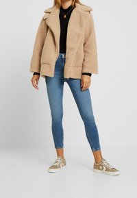 Beige faux fur jakke med reverskrave, sort sweater, blå skinny jeans og beige sneakers med hvid stjerne detaljer.