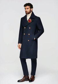 Manteau en laine bleu marine à double boutonnage avec des boutons dorés, porté sur un col roulé crème, orné d'une broche rouge en forme de cœur sur le revers.