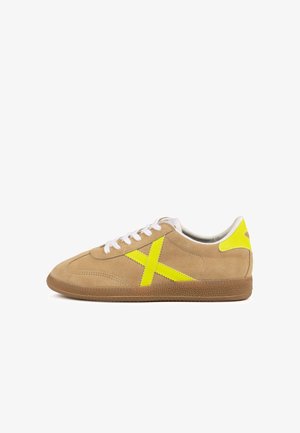 Sneaker beige in pelle scamosciata con lacci bianchi, tallone e logo giallo brillante, e suola in gomma marrone testurizzata, vista laterale su sfondo bianco.