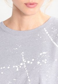 Šedá mikina s kulatým výstřhem, s designem rozstřikované barvy v bílé barvě, která má měkkou texturu a volný střih.