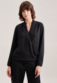 Schwarze Wickelbluse aus glattem Stoff mit langen Ärmeln, einem Knopfdetail am Ausschnitt und lockerem Schnitt. Kombiniert mit schwarzen Hosen.