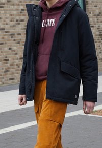 Parka negra aislada con capucha, con cremallera frontal y bolsillos laterales, llevada sobre una sudadera burdeos y pantalones de cargo naranjas.