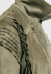 Veste en faux daim vert olive avec un col haut, fermeture éclair et un détail froncé le long de l'épaule gauche pour ajouter de la texture.