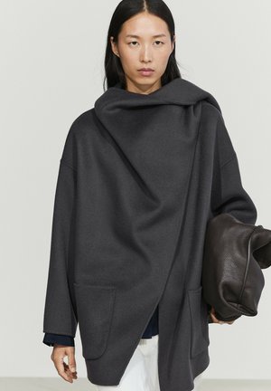 Woman with long dark hair wearing an oversized dark gray coat holding a large black textured leather bag against a plain background.