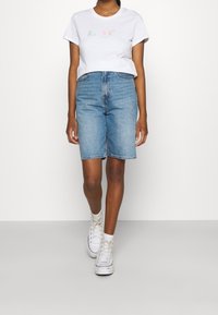 Valkoinen lyhythihainen T-paita, jossa on pastellinvärinen graafinen teksti, yhdistettynä vaaleansinisiin farkkuinbermuda-shortsseihin ja valkoisiin korkeavartisiin tennareihin.