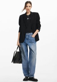 Svart texturerad blazer över en svart topp, parat med lösa blå denimjeans. Accessoarer inkluderar en svart perforerad väska och svarta skor.