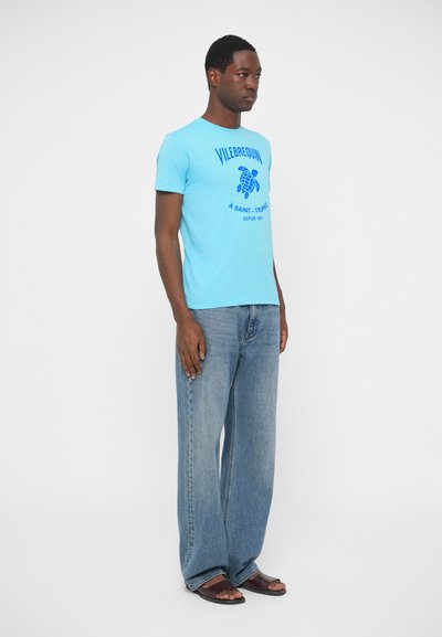 Moški v svetlobnem modrem grafičnem majici, širokih izbledelih kavbojkah in rjavih sandalih, stoji pred belo ozadjem.