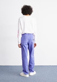 Purple cargo pants with multiple pockets and stitched accents, paired with a long-sleeve white shirt. Clear texture and casual design.