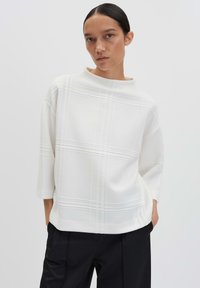 Donna con capelli scuri raccolti in un basso chignon, che indossa un maglione bianco a trama larga e pantaloni neri, in piedi con le mani in tasca contro uno sfondo neutro.