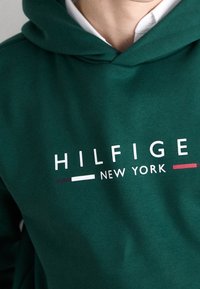 Felpa verde scuro realizzata in tessuto morbido, con il testo bianco "HILFIGER NEW YORK" e colori accentuati in borgogna e bianco, che mettono in risalto il design del logo.