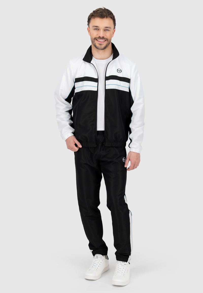 Trainingsjack en broek in zwart en wit met gestreepte accenten. Gevoerde jas met een staande kraag. Model draagt witte sneakers.