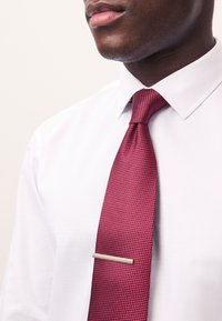 Red patterned tie with a textured fabric, featuring a straight silver tie clip, paired with a white collared shirt.