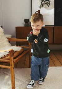 Czarna bluza z kolorowymi łatkami w kształcie uśmiechów, zestawiona z szerokimi niebieskimi spodniami cargo z denimu i czarnymi trampkami. W tle drewniane krzesło.